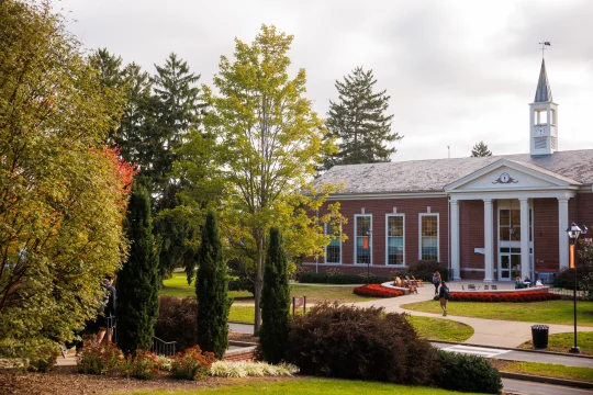 A brick building with white columns and a clock tower surrounded by green trees and landscaping. A few people walk on the path in front of the building under a partly cloudy sky.