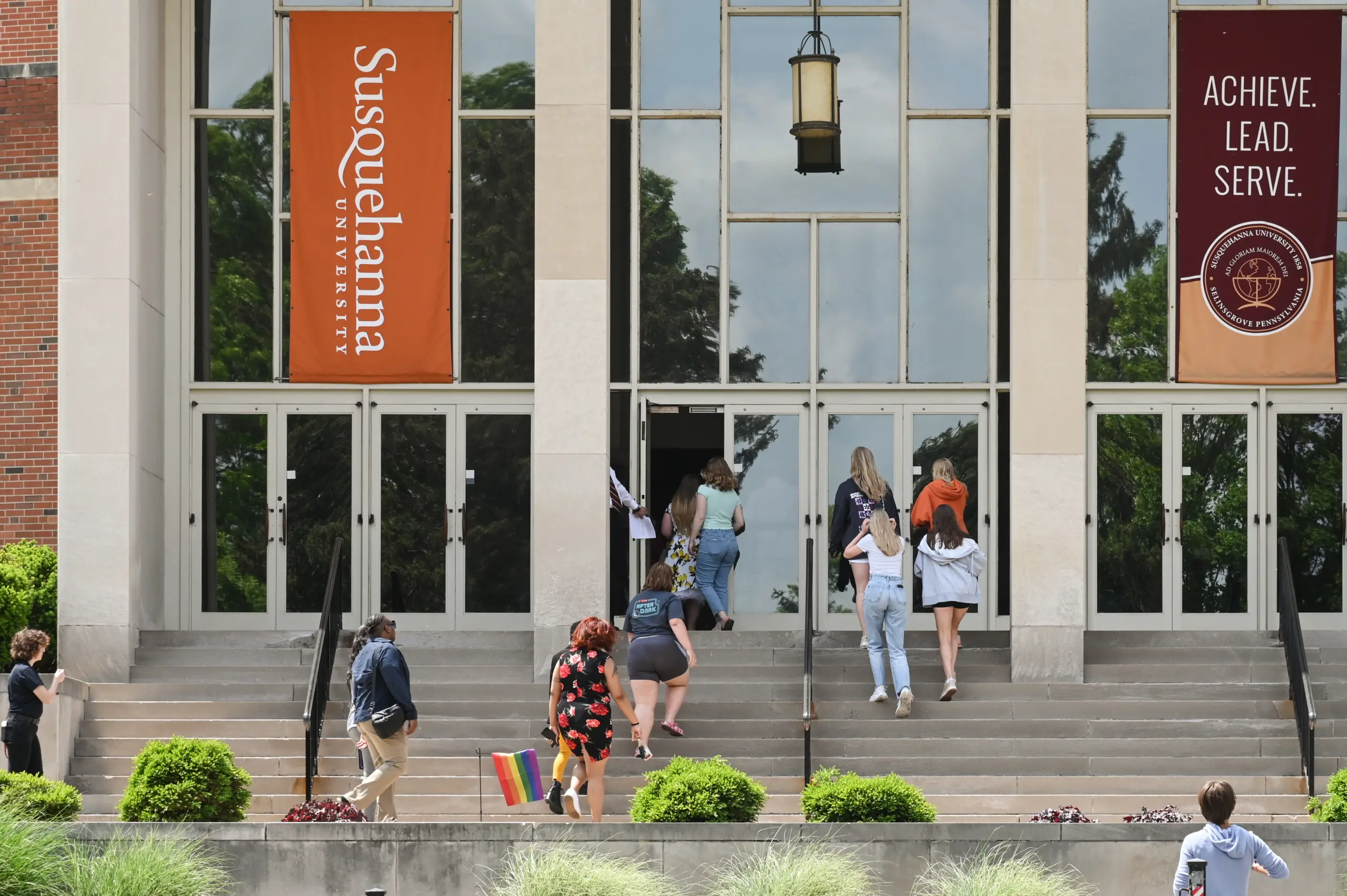 Students walk up the steps toward a building with large glass doors at Susquehanna University, where banners read “Susquehanna University” and “Achieve. Lead. Serve.” A student carries a small rainbow flag, reflecting the university’s inclusive academic community.