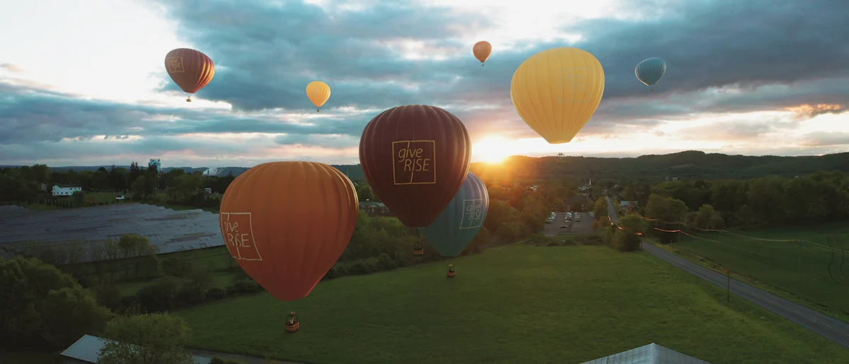 Hot air balloons of various colors float over a green landscape at sunset, with a cloudy sky and sun setting behind hills in the background.