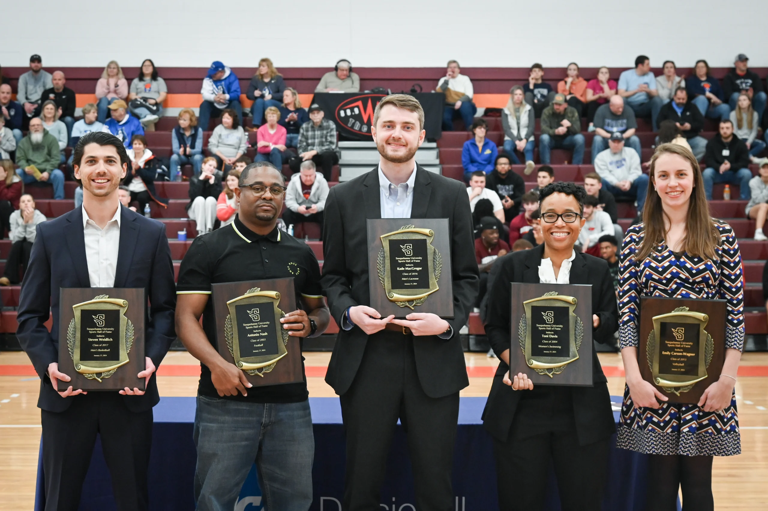 Five individuals stand in a row holding plaques at an indoor event, with an audience seated in the bleachers behind them.