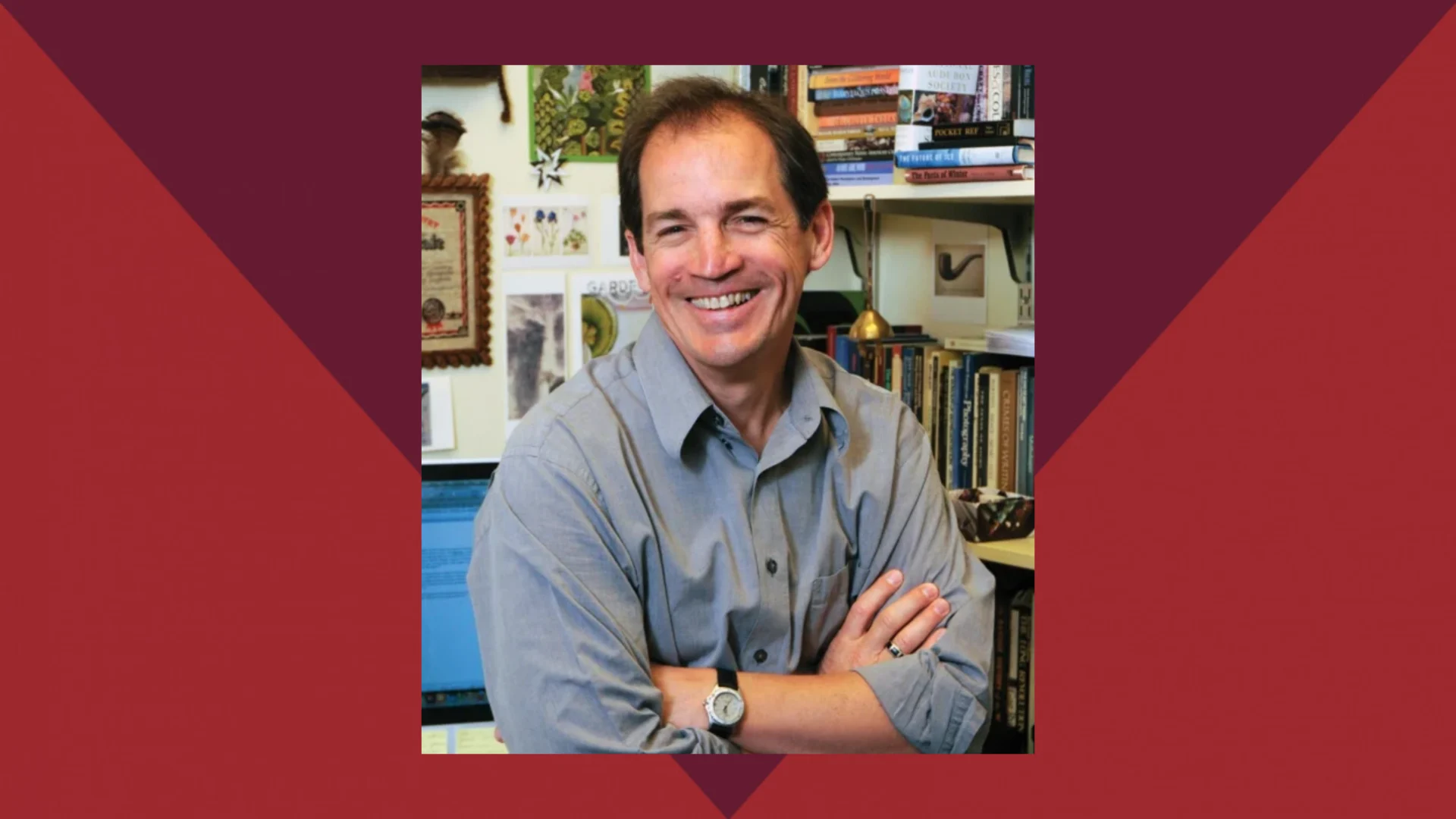A man in a gray shirt sits with arms crossed, smiling, in an office filled with books and papers.