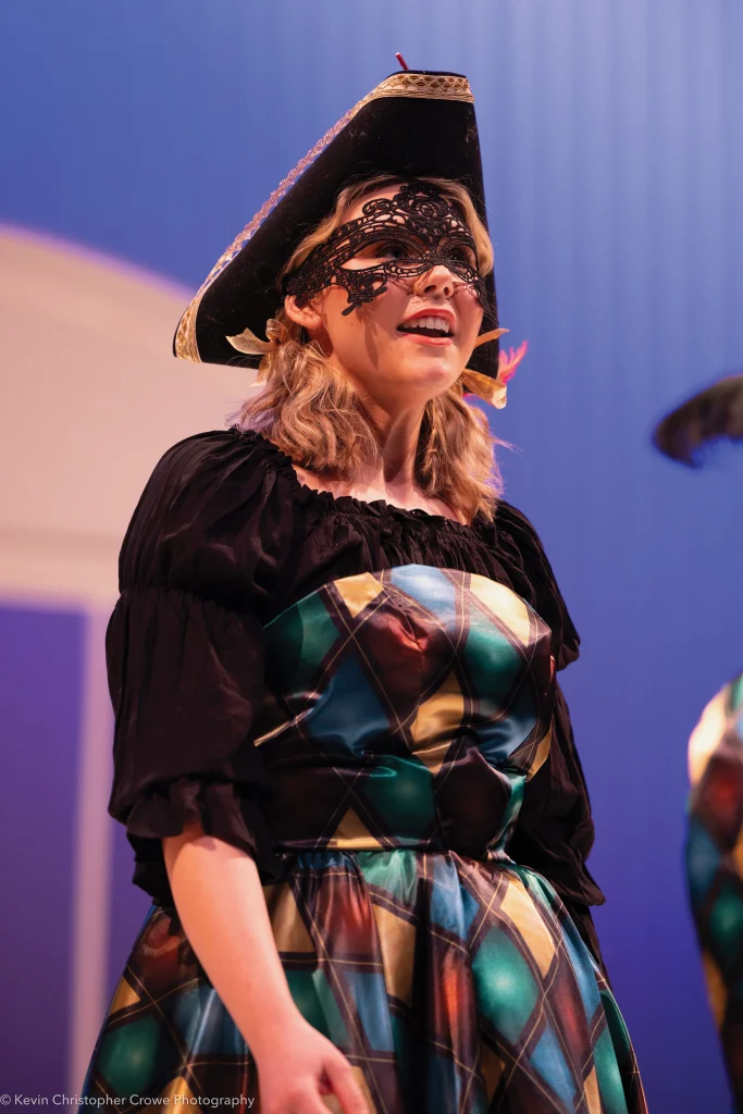 A person in a colorful harlequin costume and black masquerade mask stands on stage, wearing a tricorn hat.