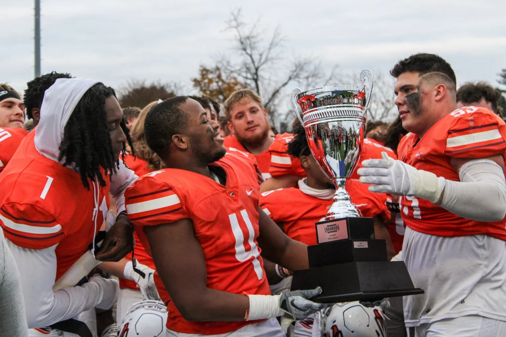 Football team players in orange uniforms celebrate, holding a large silver trophy. One player in the center smiles while displaying the trophy to his teammates. Trees and sky are in the background.