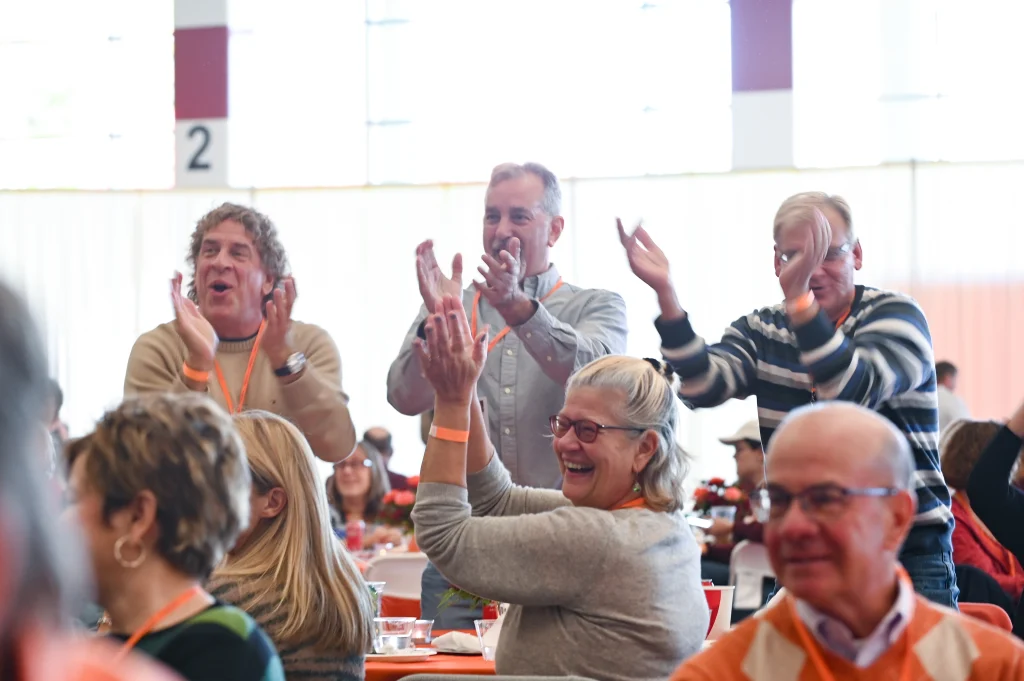 A group of people enthusiastically clapping while sitting and standing in a large indoor gathering.