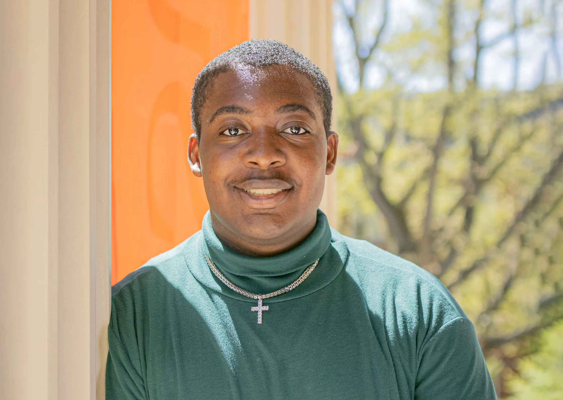 Person in a green turtleneck and cross necklace, leaning against a wall with an orange background, standing outdoors on a sunny day with trees in the background.