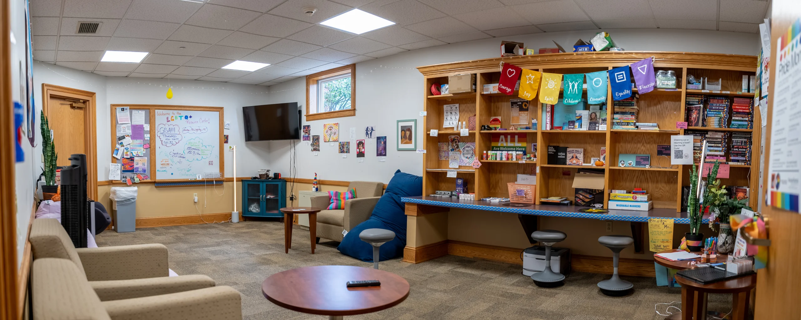 A cozy room with chairs, a small table, a wall-mounted TV, shelves of books, and colorful flags. A whiteboard with drawings and notes is on the left.