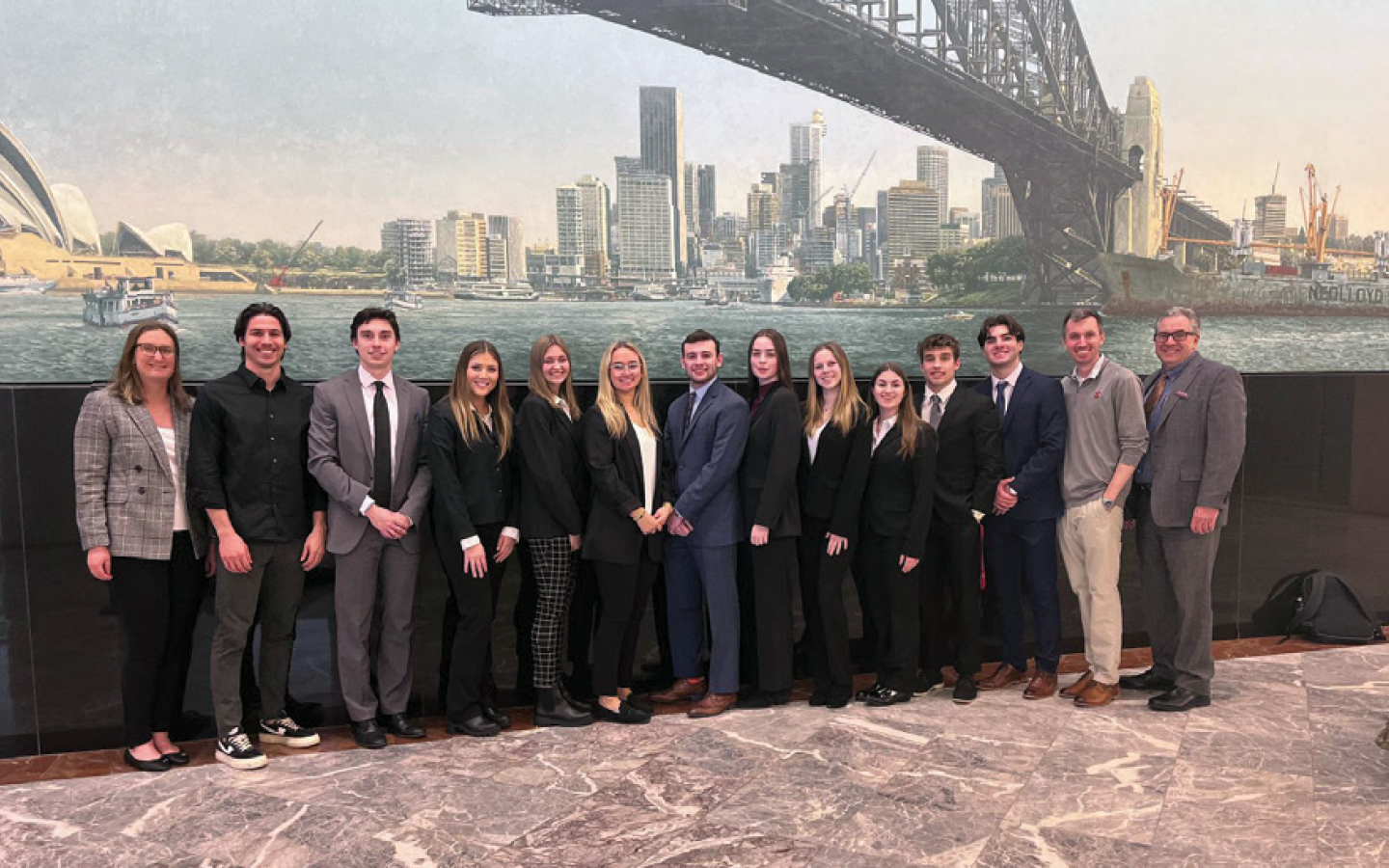 A group of people in formal attire stand in front of a large wall mural depicting a cityscape with a bridge and skyline.