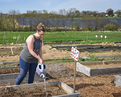 A woman waters a garden bed with a watering can in a rural setting. She's wearing a tank top and leggings. A sign reading "bee buzz!" is planted in the garden. Fields and trees are visible in the background under a clear sky.