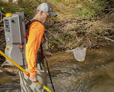 A person wearing waders and an orange long-sleeve shirt stands in a shallow stream with a small net. They have a large backpack and wear a cap, surrounded by foliage and branches.