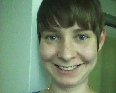A person with short brown hair smiles at the camera. The background is a plain indoors setting with a white wall.