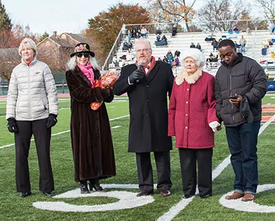 Five people stand on a football field. Four are dressed in winter coats and one is using a smartphone. The stands in the background have a few spectators. Trees with autumn leaves are visible.