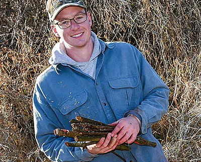 A person wearing glasses, a cap, and a blue button-up shirt smiles while holding a bundle of sticks. They are standing outdoors in front of dry grass and bushes.