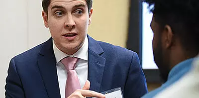 A man in a blue suit and pink tie speaks to another person, gesturing with his hand. The man is wearing a name tag, and the conversation appears to be taking place indoors.