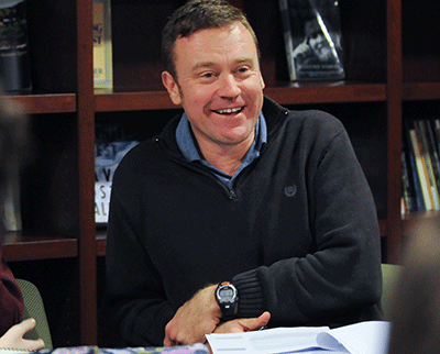 A man wearing a dark sweater and a wristwatch is sitting at a table with papers, smiling. Shelves with books are visible in the background.
