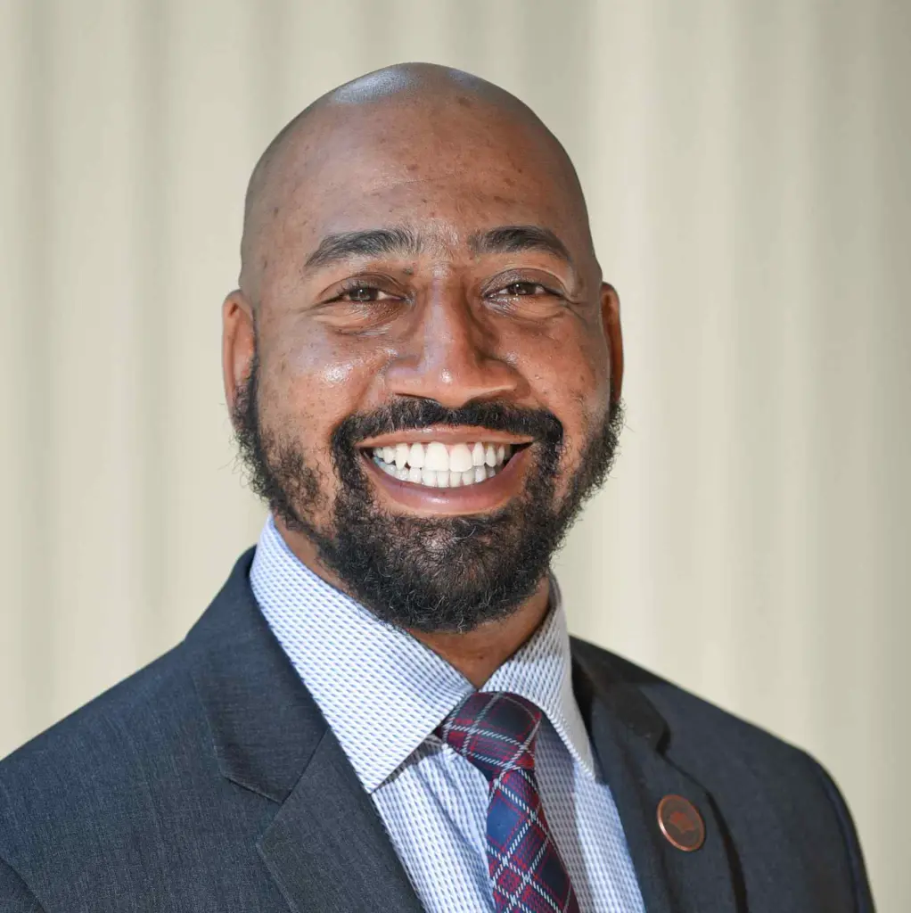 A smiling man with a beard wearing a gray suit, white shirt, and patterned tie stands against a plain light-colored background.