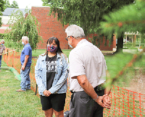 Two people wearing masks are talking in a grassy area next to a red mesh fence. A man in a blue shirt stands in the background. Trees and a brick building are visible.