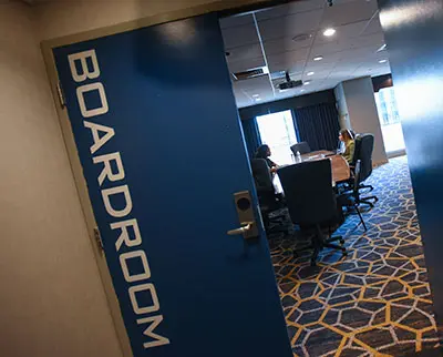Open door to a boardroom with a large conference table and chairs. Two people are seated at the table. The blue door has "BOARDROOM" in large white letters. The carpet features a geometric pattern. Natural light streams in from windows.