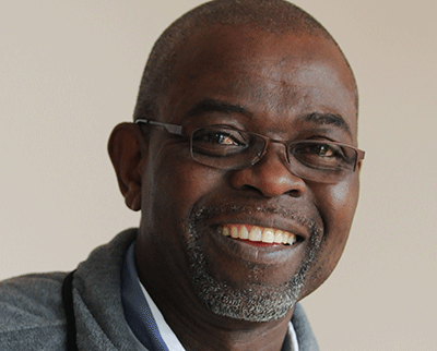 A smiling man wearing glasses and a gray sweater over a blue and white shirt. The background is plain and light-colored.