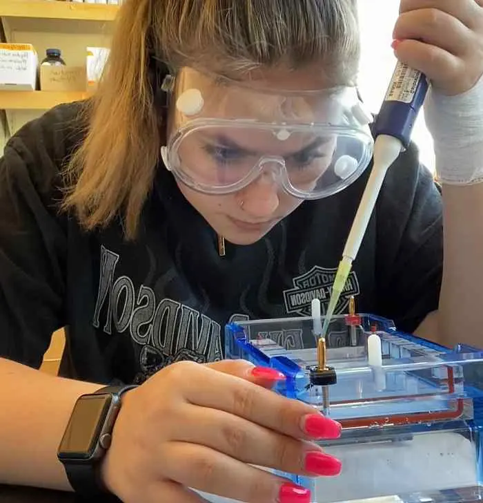 A person wearing protective goggles is focused on using a pipette for a science experiment. They are handling a rectangular container and wearing a black shirt and smartwatch. Their nails are painted bright pink.