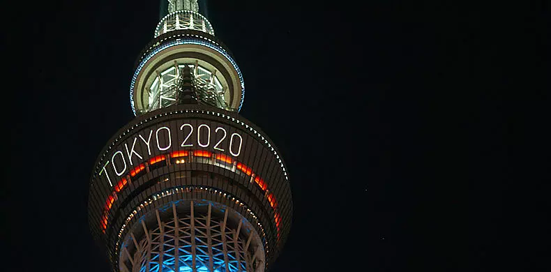 Tokyo Skytree tower illuminated at night, displaying the words "TOKYO 2020" in bright white lights, referencing the Tokyo 2020 Olympic Games.