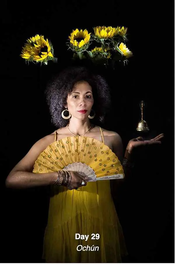 A woman in a yellow dress holds a decorative fan and a small bell. Sunflowers float above her head. Text below reads "Day 29 OchĂşn." The background is dark, highlighting the vibrant colors.