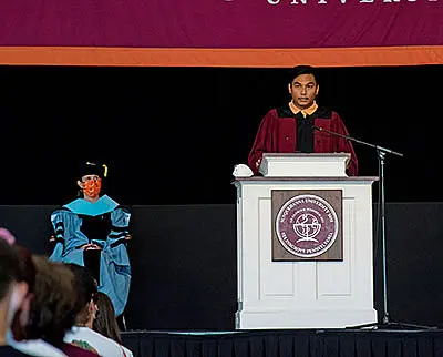A person in maroon academic robes speaks at a podium on stage, with a university emblem. An individual in blue academic regalia sits nearby. The setting is a formal academic event, with an audience visible in the foreground.