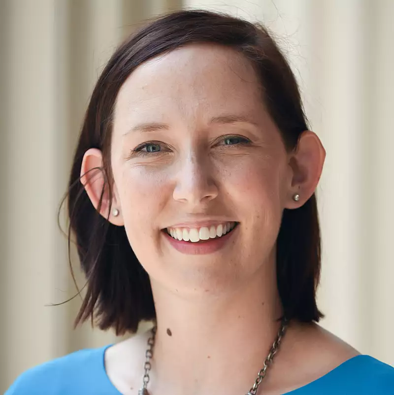A woman with short brown hair, wearing a blue top and a necklace, smiles at the camera. The background is softly blurred with vertical beige lines.