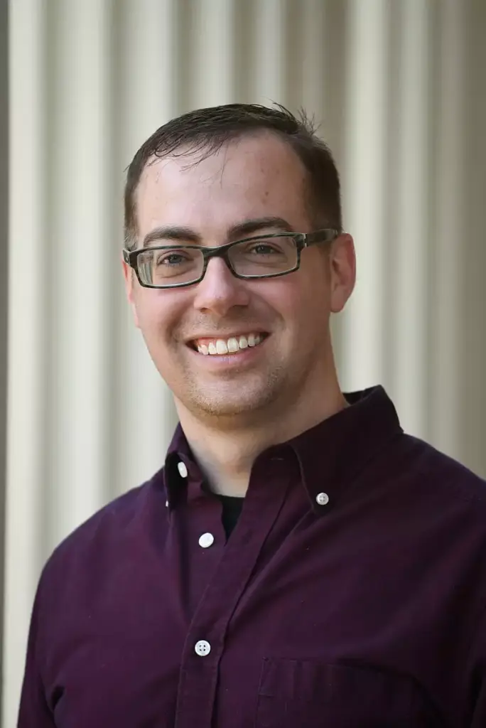 A man with short brown hair and glasses smiles at the camera. He is wearing a maroon button-up shirt over a black T-shirt, standing in front of a light-colored, blurred background with vertical lines.