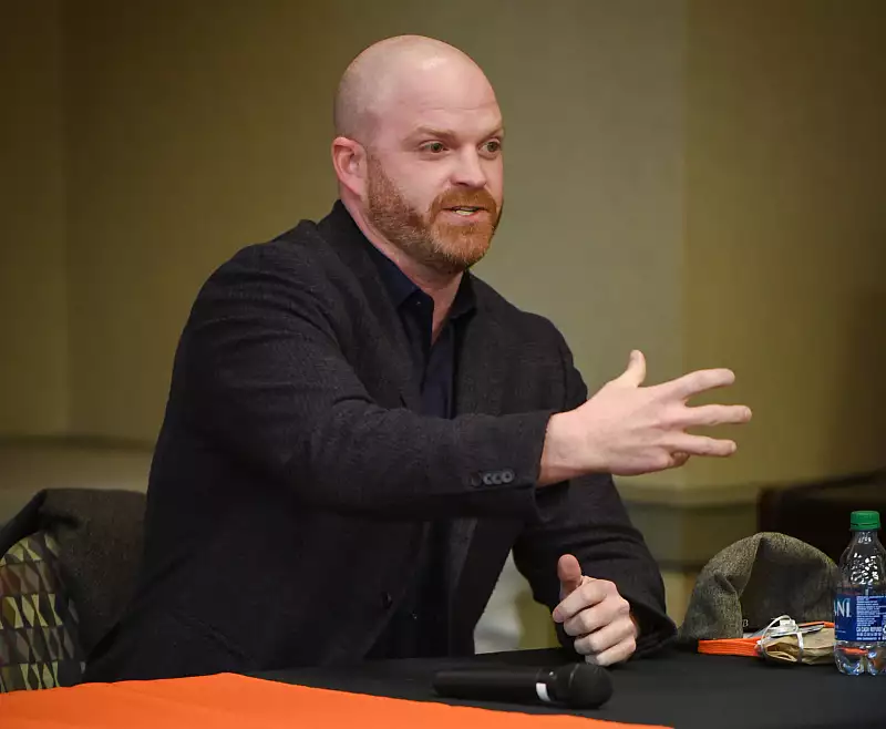 A bald man with a beard sits at a table, gesturing with his right hand while speaking. He wears a dark blazer and shirt. A microphone, glasses, and a water bottle are on the table in front of him.