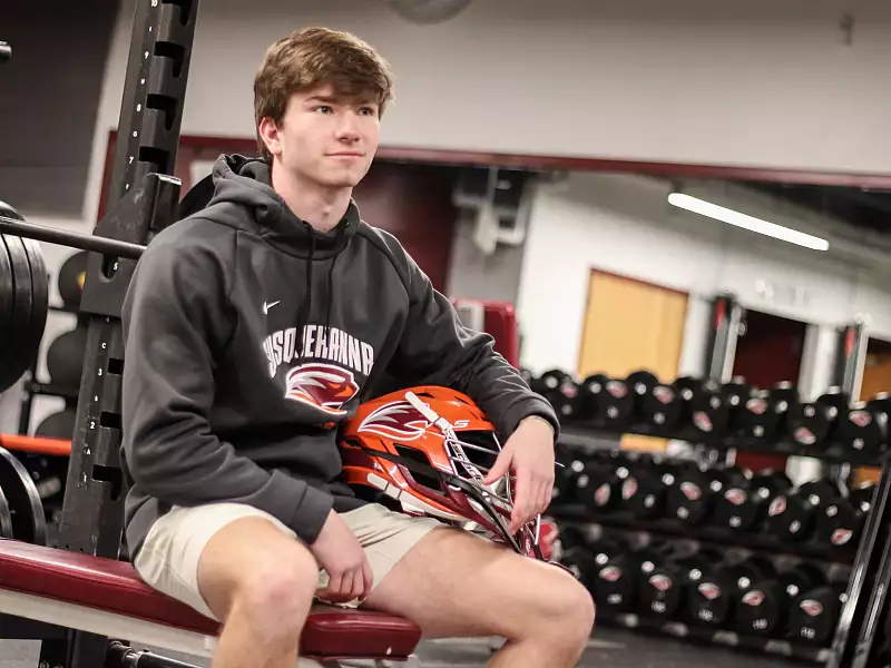 A young man sits on a bench in a gym, holding a red lacrosse helmet. He is wearing a dark hoodie with "Susquehanna" printed on it and beige shorts. The background shows a weight rack and several dumbbells.
