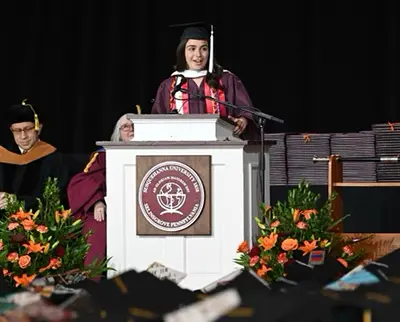 A student in graduation attire speaks at a podium on stage. The podium displays a university emblem. Faculty members sit behind her, and stacks of diplomas and floral arrangements are visible. Graduates' mortarboards can be seen in the audience.