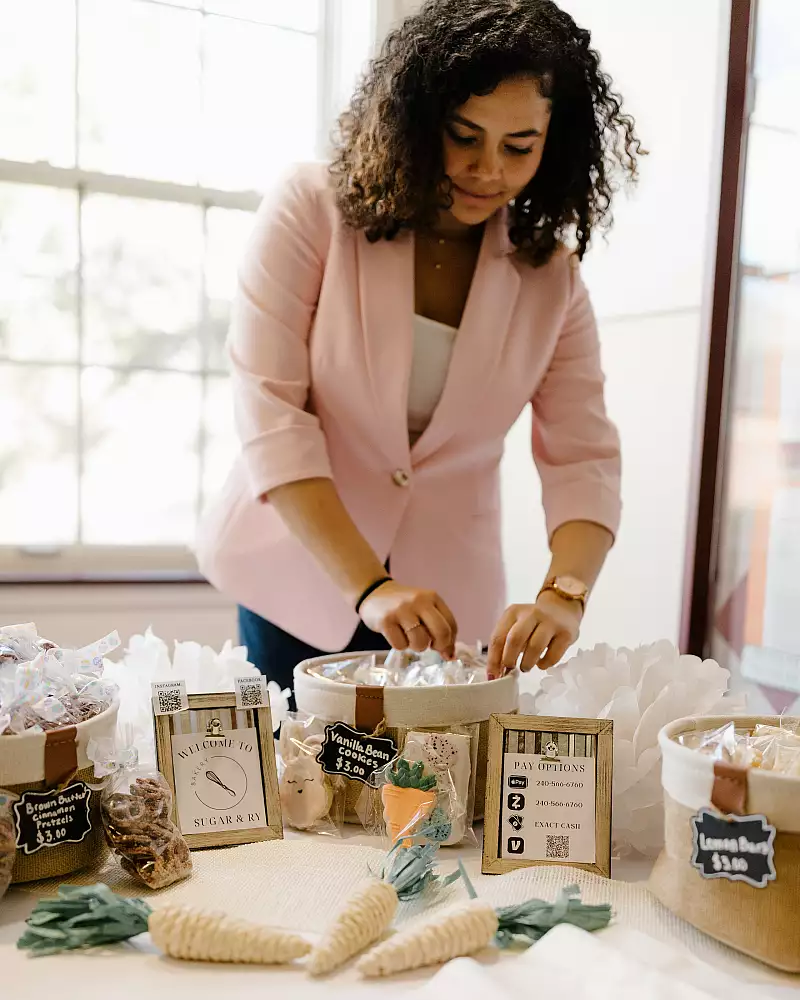 A woman in a pink blazer arranges items on a table filled with baskets of small wrapped goods. Signs with prices are displayed, and soft natural light filters through a window in the background.