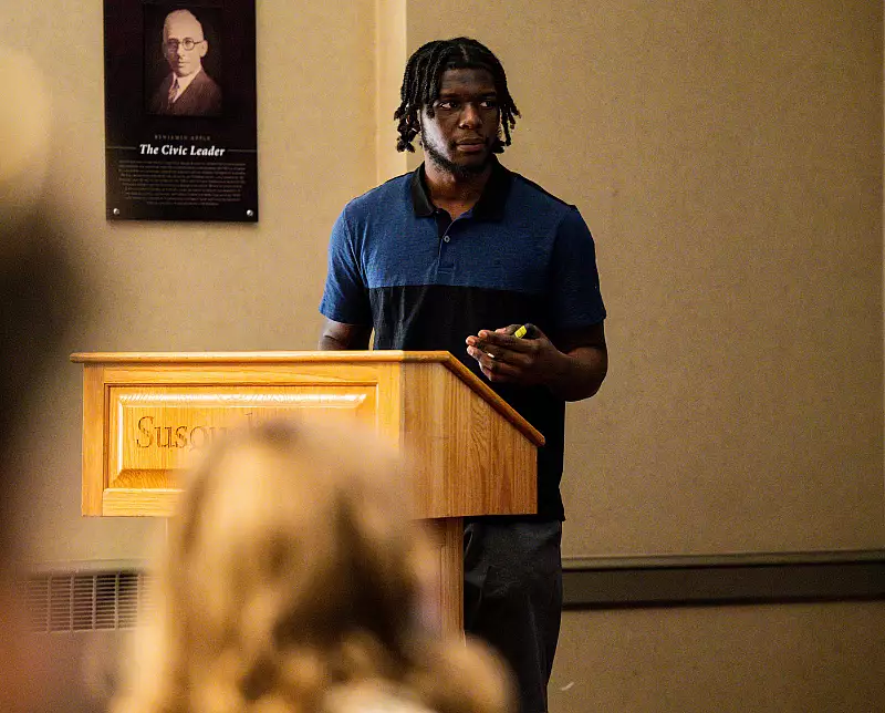 A person with dreadlocks, wearing a blue and black polo shirt, stands behind a wooden podium labeled "Susquehanna." They are holding something small, speaking to an audience. A poster on the wall in the background features another person.