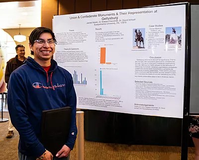 A person stands in front of a research poster titled "Union & Confederate Monuments & Their Representation at Gettysburg." The poster includes text, graphs, and images. The person is smiling and holding a folder.