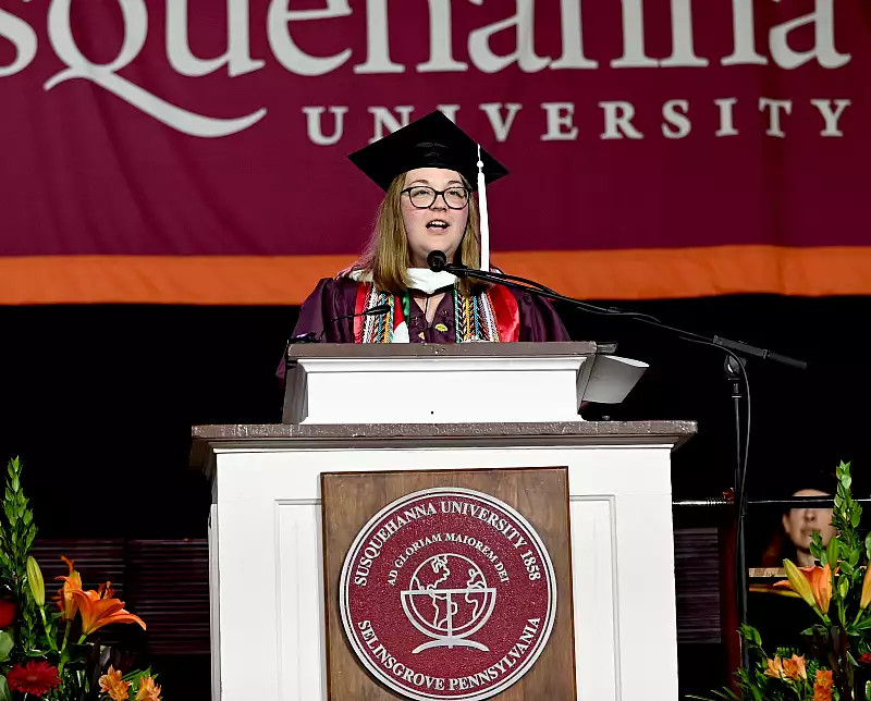 A graduate in cap and gown speaks at a podium decorated with Susquehanna University signage. The background features a banner with the university's name. Flower arrangements are visible by the podium.