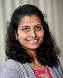 Smiling woman with long curly black hair, wearing a pink top and gray sweater, standing against a light gray background.