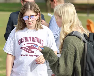 Two people are outdoors. One wears a Susquehanna River Hawks T-shirt and sunglasses, looking at something. The other has long hair and a green sweater, holding a pen and paper. Blurred people in the background suggest a campus or park setting.