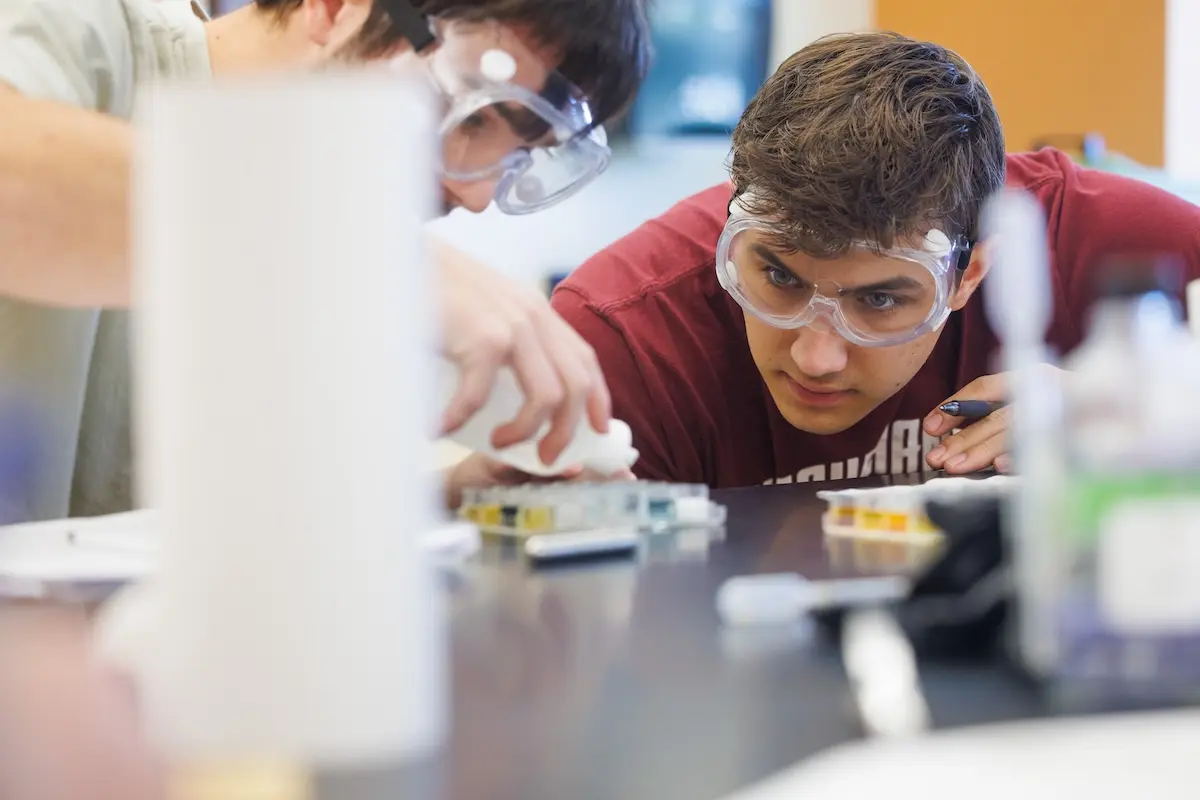Two students wearing safety goggles work intently on a science experiment at a lab table. One student carefully drops liquid into a container, while the other observes closely. Various lab equipment is scattered across the table.
