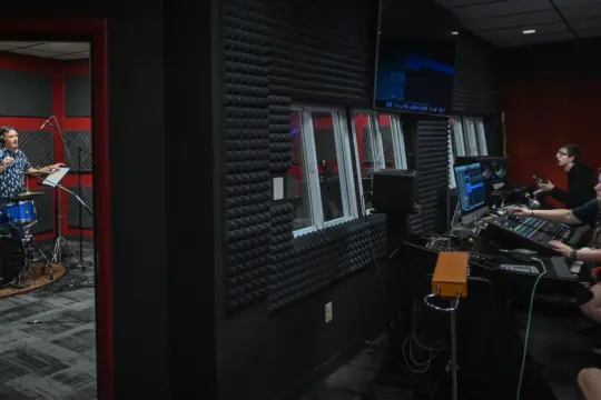 A recording studio with a drummer playing in a soundproof room. Two people are inside the control room, one operating a soundboard and the other watching computer screens. The walls are covered with acoustic foam panels.