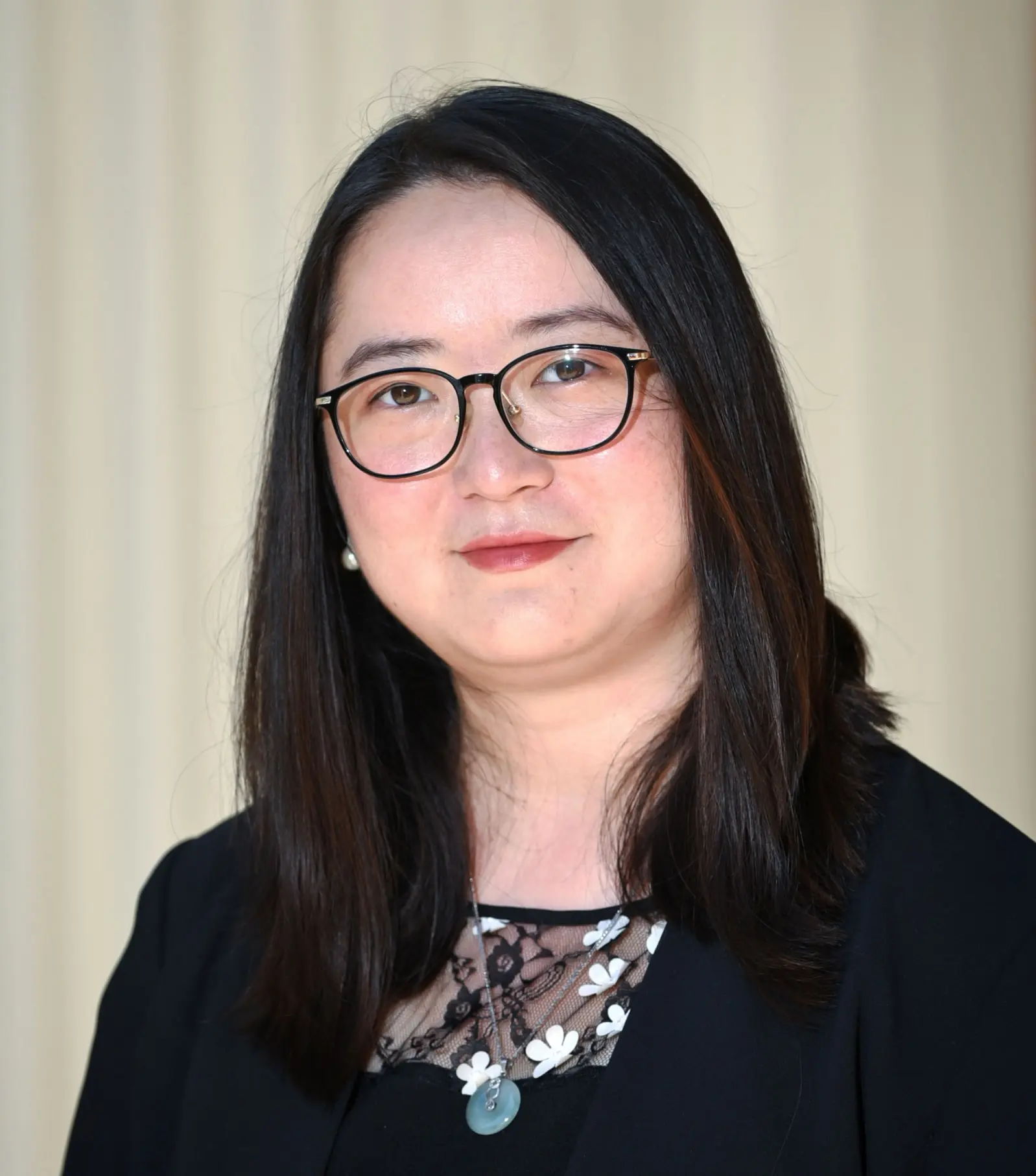 A person with long, dark hair and glasses is wearing a black top with a floral design at the neckline. The background is a light, neutral color.