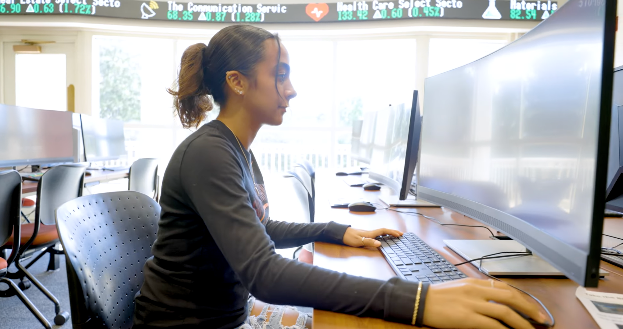 A person sits at a desk in an office setting, using a widescreen computer to analyze accounting data. Stock market ticker displays are visible in the background. The room is furnished with multiple chairs and desks, while large windows bathe the space in natural light.