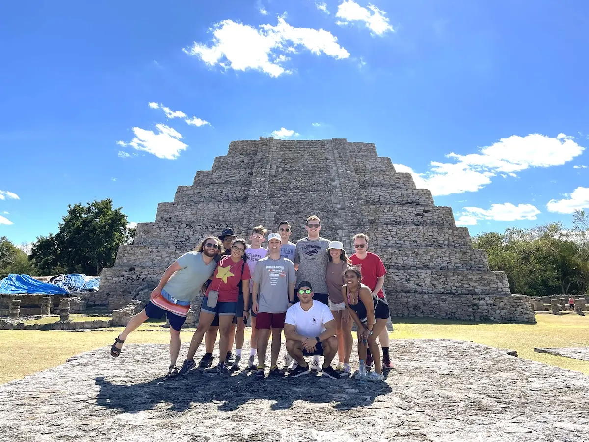 A group of people posing in front of a large stone pyramid under a clear blue sky. Some are wearing hats and sunglasses, and all are casually dressed. They stand on a stone platform with trees in the background.