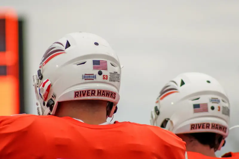 Two American football players in orange jerseys and white helmets with "River Hawks" labels stand on the field, seen from behind. A blurry scoreboard is visible in the background.