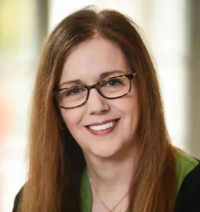 A woman with long brown hair and glasses smiles at the camera. She is wearing a dark top with a green collar. The background is softly blurred, creating a warm, natural atmosphere.