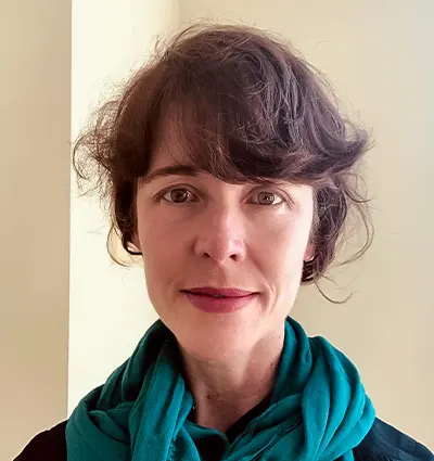 A person with short, wavy brown hair and a teal scarf stands in front of a neutral background.