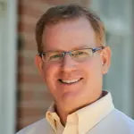 A man with short hair and glasses is smiling in an outdoor setting. He is wearing a light-colored button-up shirt. The background features a brick wall and a window.