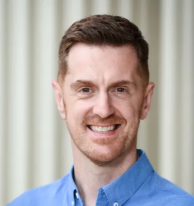 Smiling person with short brown hair, wearing a blue button-up shirt, stands in front of a blurred neutral background.