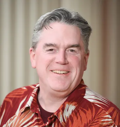 A man smiling while wearing a red and white patterned shirt. He has short gray hair and is standing against a plain, blurred background.