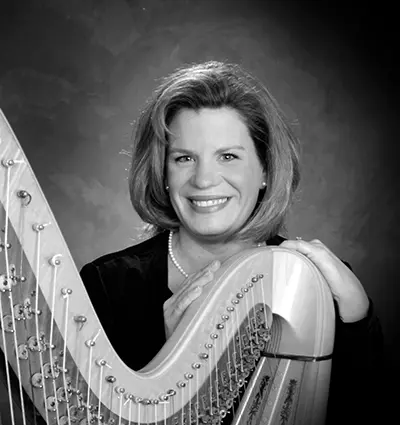 A black and white portrait of a person smiling, standing beside a harp. They have shoulder-length hair and are wearing a dark top. Their hands are gently resting on the harp. The background is softly textured.