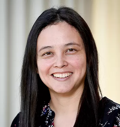 A person with long black hair is smiling, wearing a black top with floral patterns. The background is softly blurred in neutral tones.